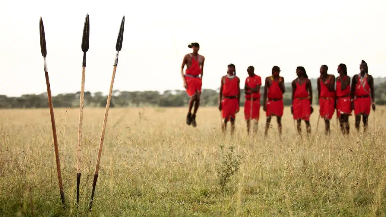 7-Days-in-Kenya-That-Will-Completely-Change-Your-Life-1280x720 Maasai warriors in traditional red shuka perform the adumu jumping dance in the Kenyan savanna, with spears planted in the grass in the foreground.