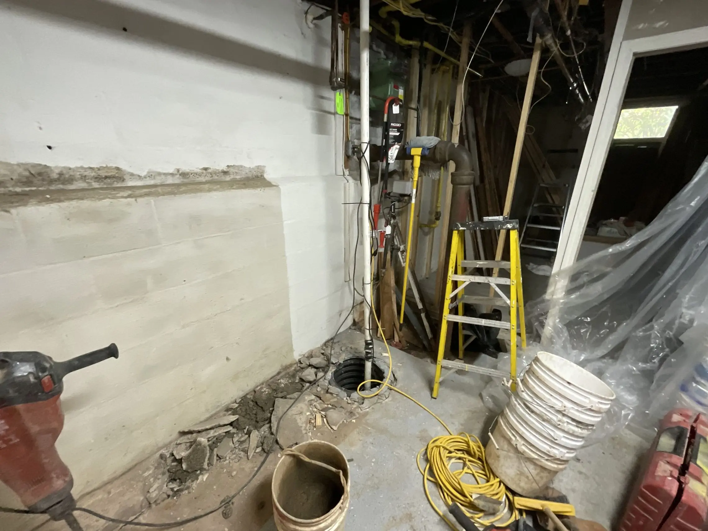 A basement corner under construction with exposed pipes and wires. Yellow ladder, power tools, and buckets scattered around. The scene feels industrial and busy.