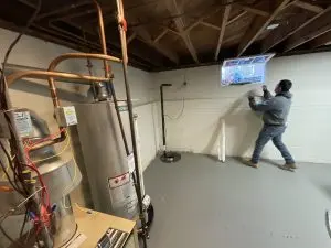 A person in a gray hoodie adjusts a small window in a clean, unfinished basement. Exposed ceiling beams and a water heater are visible. Functional atmosphere.