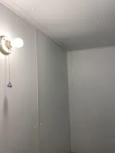 A small, minimalist room with textured white walls and a ceiling. A single exposed light bulb emits soft light, creating a simple, unadorned atmosphere.