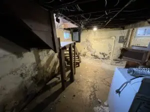 Dimly lit basement with exposed wooden beams and a small window. Worn walls show signs of peeling paint and water damage. A staircase leads up, while discarded objects and debris litter the floor, creating an abandoned and neglected atmosphere.