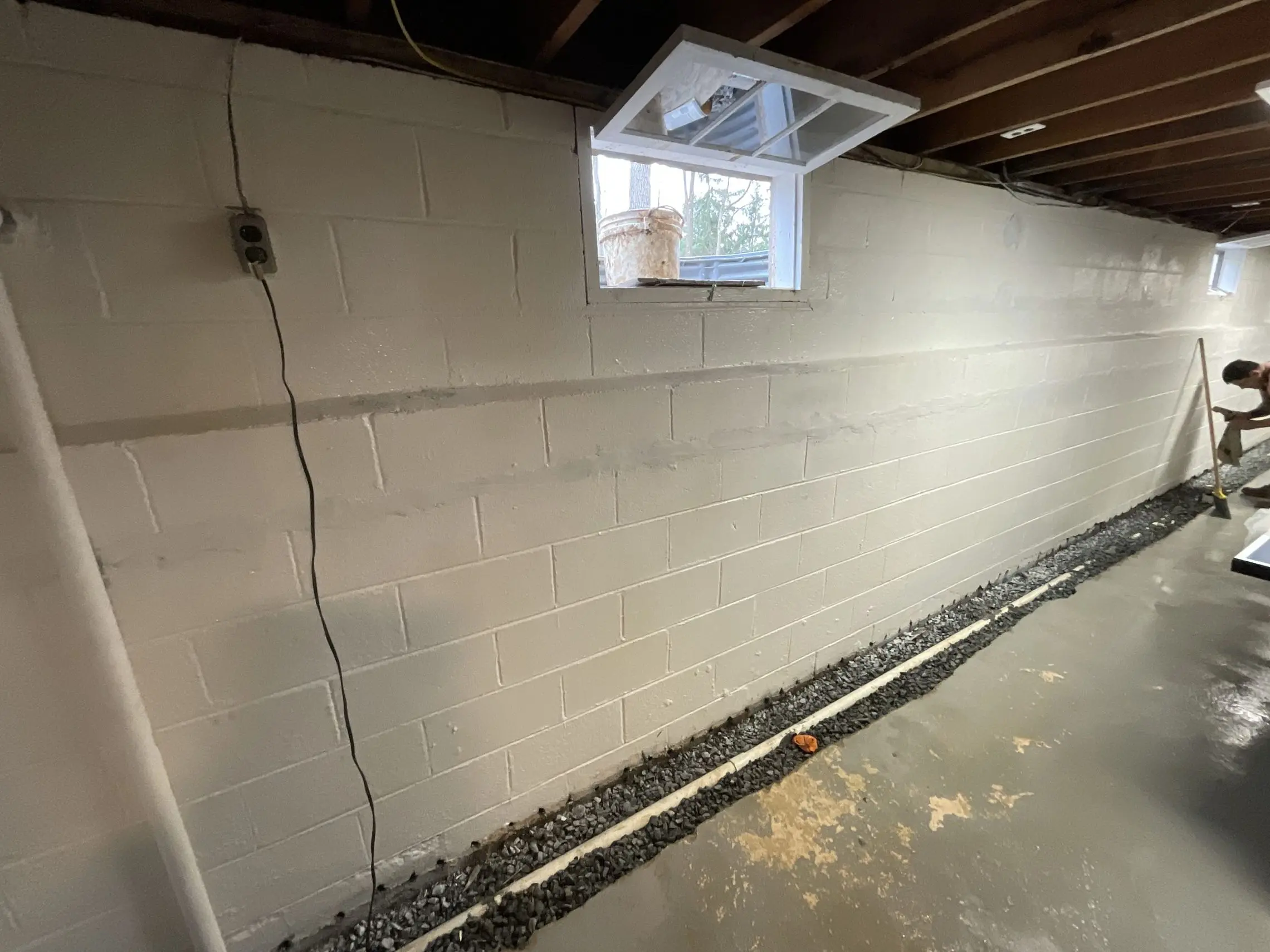 A basement with exposed beams shows a white-painted cinderblock wall. An open window lets in light. A trench with gravel runs along the floor, and a person working with a tool is visible at the far end.