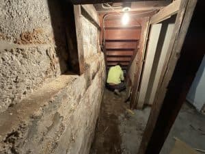 A person in a bright hoodie works under dim light in a cramped, old basement with rough concrete walls and wooden stairs, creating an eerie atmosphere.