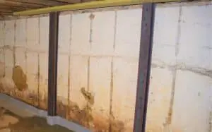 A basement wall with visible signs of water damage, stains, and cracks is reinforced with metal beams, conveying an atmosphere of repair and restoration.