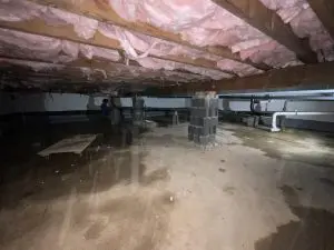 Crawl space with exposed pink insulation under wooden beams. Damp floor with pipes visible. Worker in the background suggests repair work. Gloomy atmosphere.