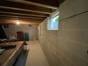 A dimly lit basement with exposed wooden beams and cinder block walls. A small window lets in natural light. Tables hold scattered items, creating a cluttered feel.