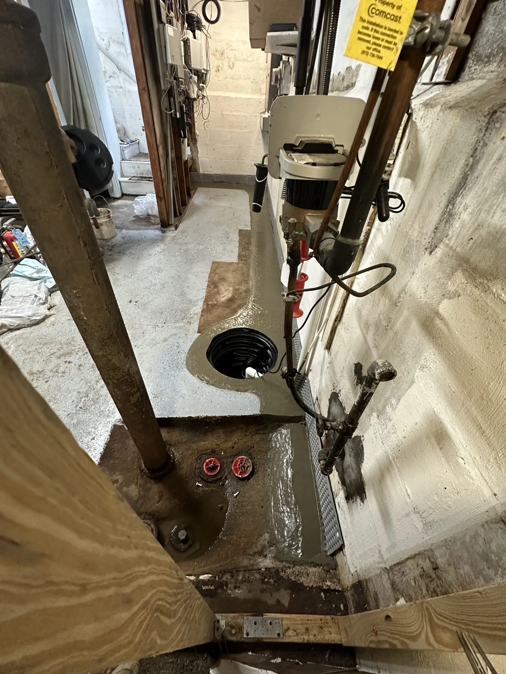 Basement corner with concrete floor, wall pipes, and an open sump pit. Fresh concrete surrounds the pit, suggesting recent work. The space is narrow and functional.