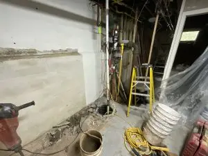 A basement corner under construction with exposed pipes and wires. Yellow ladder, power tools, and buckets scattered around. The scene feels industrial and busy.