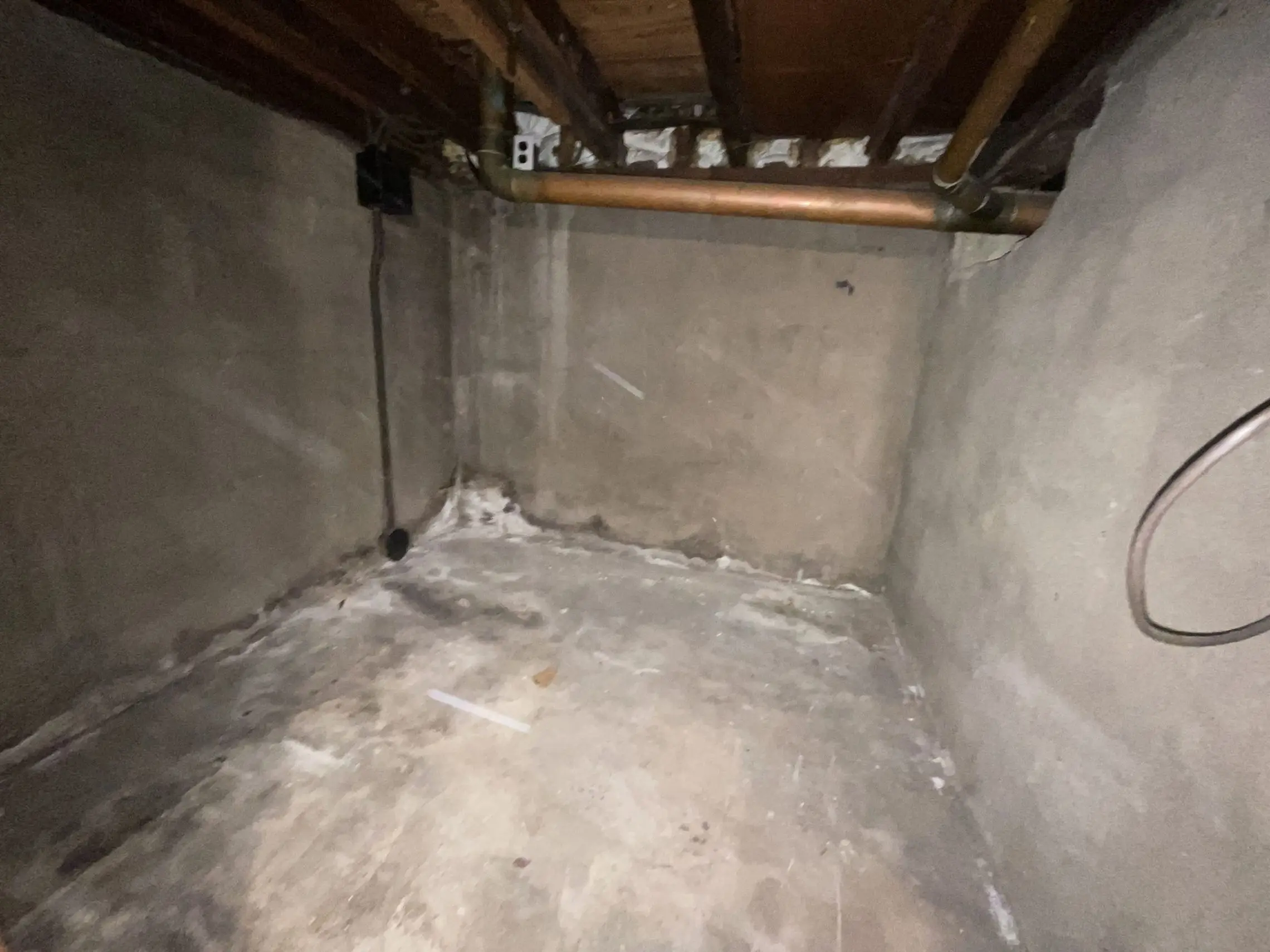 A dimly lit basement corner with unfinished concrete walls and floor, exposed wooden ceiling beams, and copper pipes. The scene feels cold and utilitarian.