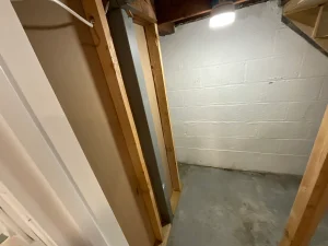 A person in a casual outfit adjusts a window in a basement with unfinished ceiling beams. A ping pong table is nearby, surrounded by various stored items.