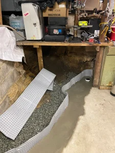 A basement under repair, showing a dirt and gravel floor with gray drainage mats partially installed. Workbench with tools and boxes behind. Construction in progress.