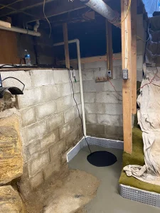 A basement corner with newly installed drainage, exposed pipes, and a sump pump. Concrete walls, unfinished ceiling, and a tarp covering steps create a work-in-progress feel.