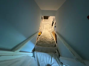 Narrow staircase with white protective covering leading downward. Walls are light blue, and a person is partially visible at the bottom. Dim lighting sets a calm tone.