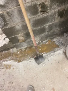 A shovel with a wooden handle leans against a rough, unfinished brick wall. The floor shows scattered debris, suggesting recent construction work.