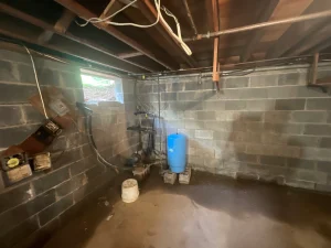 Dimly lit basement with concrete walls and exposed beams. A blue water tank is on the floor near plumbing pipes. The space feels damp and utilitarian.