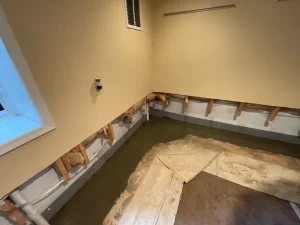 Basement corner with yellow walls, visible piping, and exposed framing. Floor is partially removed, revealing damp concrete and drainage system.