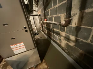 Basement scene with furnace and exposed pipes against a gray brick wall. The atmosphere is industrial, and lighting is dim, highlighting textures.