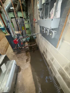 Flooded basement with water pooling on concrete floor near utility pipes and electrical conduits on walls. Tone suggests urgency and concern.