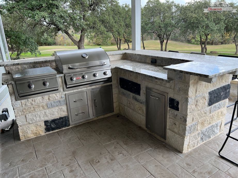 L-shaped limestone kitchen with granite bar and side burner by Diamond Decks in San Antonio, TX