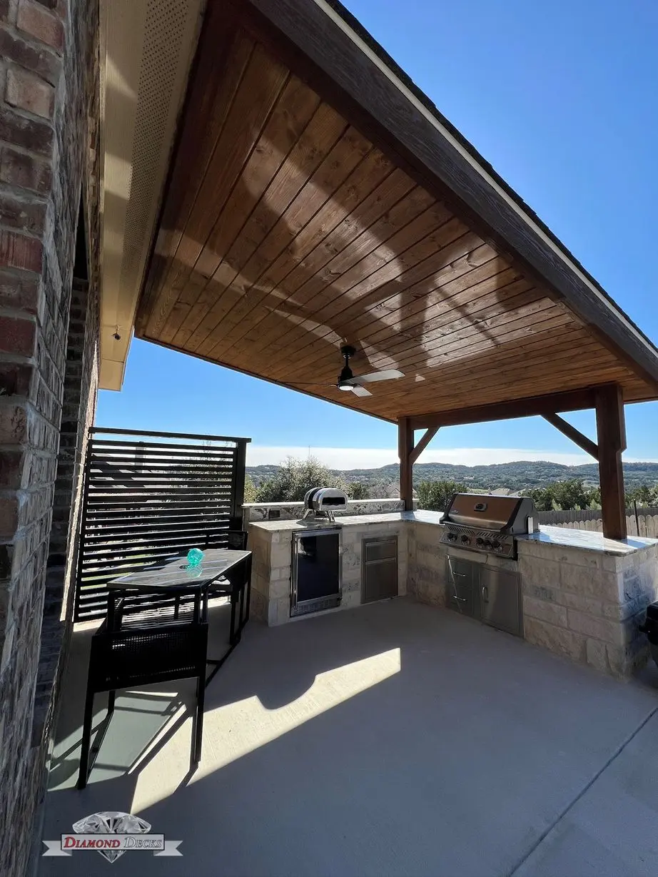 Wood patio cover shading limestone kitchen and lounge seating by Diamond Decks in San Antonio, TX