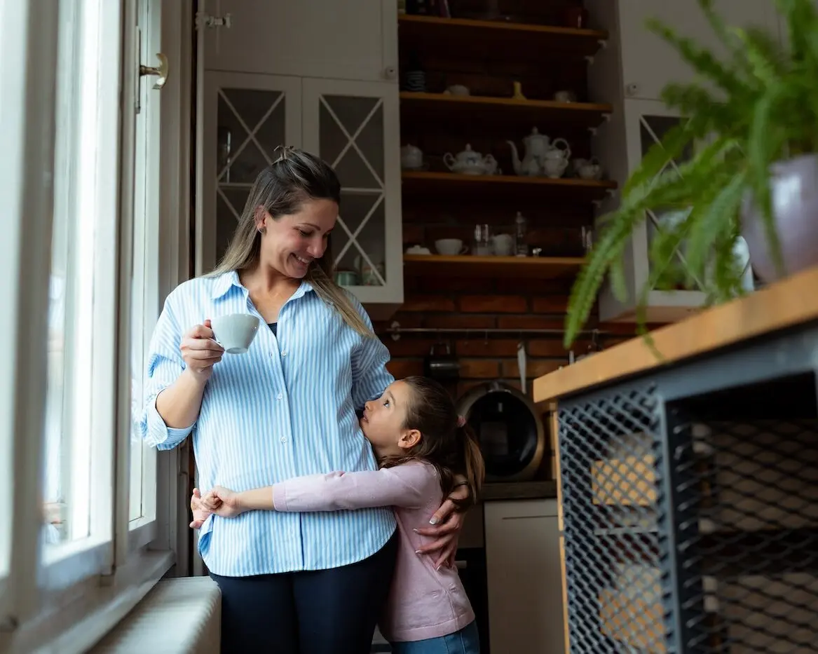 Mother enjoys a cup of coffee while her daughter hugs her affectionately in their modern kitchen