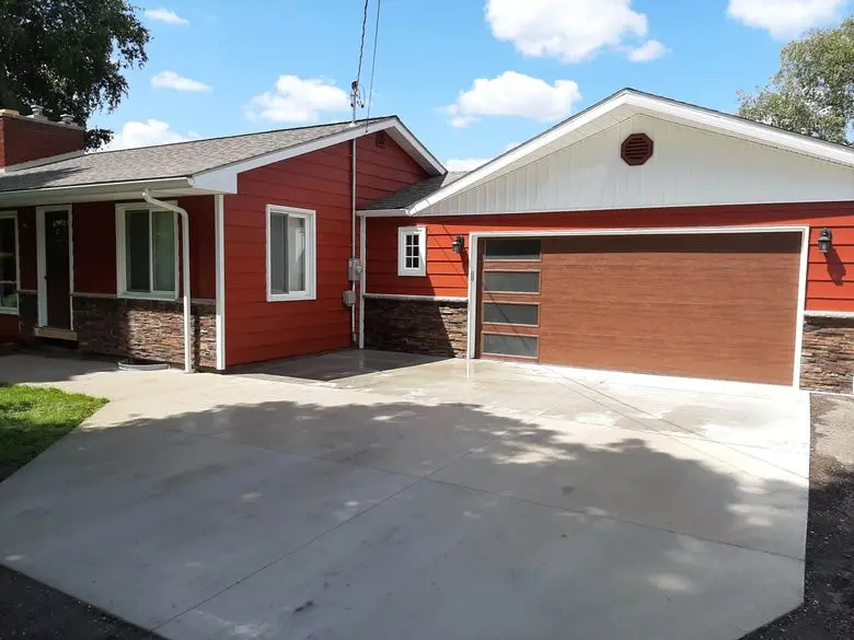 Modern red ranch-style home exterior with attached single-car garage featuring a contemporary wood-look sectional garage door and four horizontal frosted glass window inserts along the left side. White gable trim with circular attic vent, stone veneer wainscoting along lower façade, and black wall-mounted lantern lights flanking the garage. Fresh concrete driveway in foreground with wet surface reflections. Blue sky with scattered clouds and mature trees surrounding the residential property.