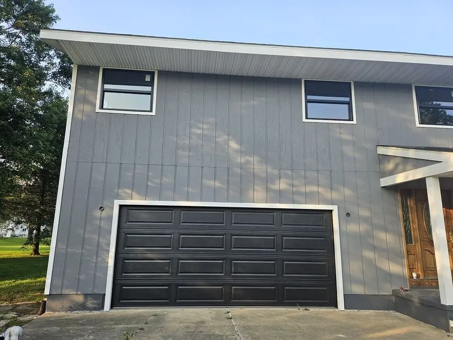 Two-story residential home exterior with gray vertical board-and-batten siding and white trim. Black raised-panel sectional garage door centered below two rectangular upper-level windows with white frames.