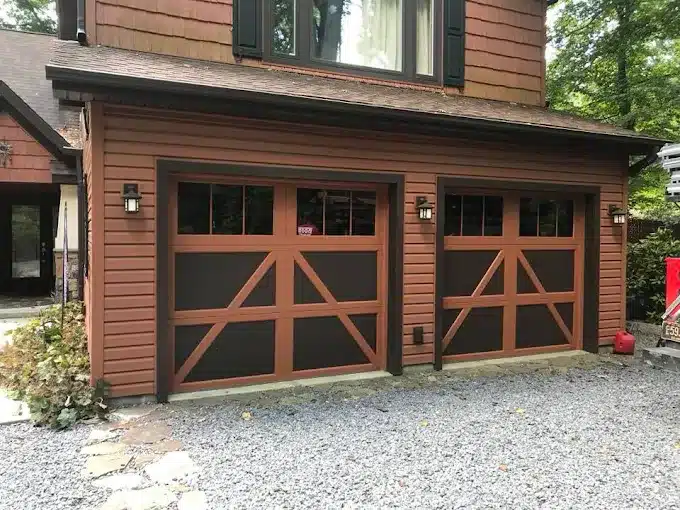 Two brown carriage-style garage doors with rustic wood-look trim and dark recessed panels on a residential home exterior. Each door features upper rectangular window panes, diagonal crossbuck design accents, and black decorative hardware. House façade includes brown horizontal siding, black shutters on upper windows, and wall-mounted lantern lights beside each garage door. Gravel driveway in foreground with stone walkway leading to side entrance.