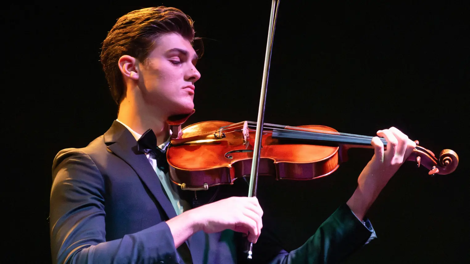Young male violinist in a dark suit and bow tie performing on stage under warm dramatic lighting against a black background
