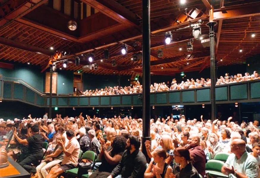 mbaw-concept1-01 Large audience filling a wood-beamed concert hall with a balcony, applauding and raising hands during a live performance.