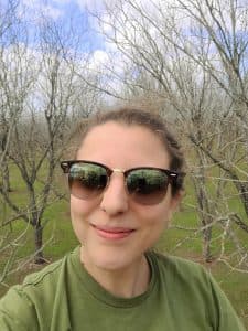 A selfie of a woman wearing sunglesses and a green shirt in a dormant pecan orchard.