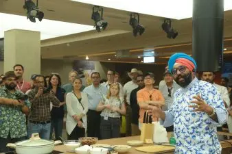 A chef speaks to a camera off to the side with a crowd of onlookers in the background.