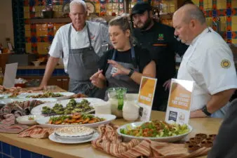 Four chefs stand behind a kitchen island covered in displayed dishes. The chef in the middle gestures toward one of the dishes, explaining what's in it.