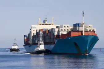 Large container ship loaded with refrigerated and standard containers approaching port, assisted by two tugs and followed by pilot cutter.