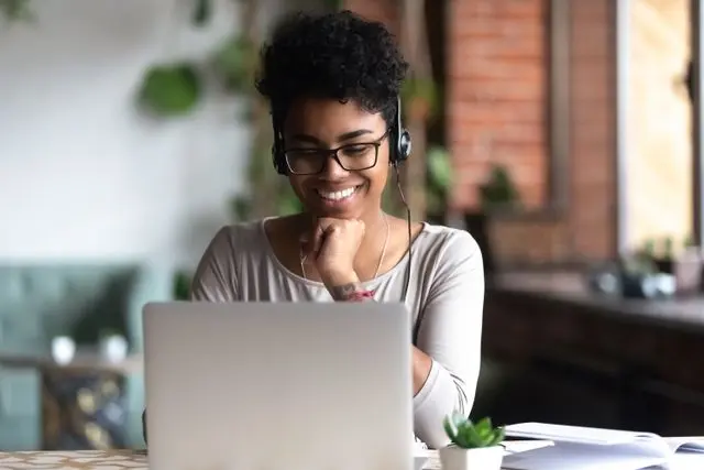 Smart student learning using internet and headphones Black woman smart student girl sitting at table in university cafe alone wearing glasses looking at computer screen using headphones listening online lecture improve language skills having good mood
