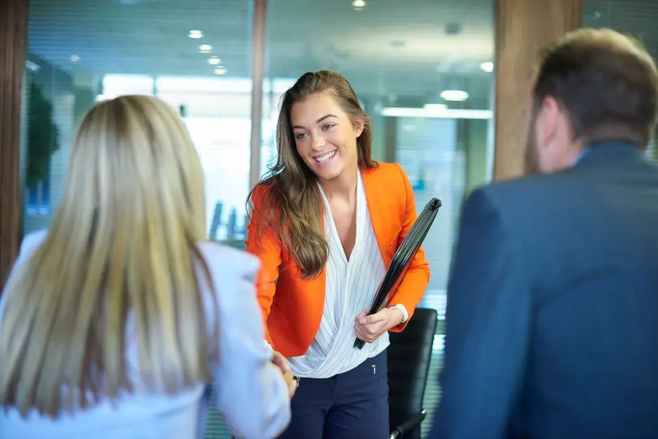 job interview first impressions a young graduate walks into an interview room full of confidence and positivity energy . She is holding her cv and smiling at the interview panel before her. She is wearing blue trousers with an orange suit jacket , as she opens the door and strides in and shakes hands with a woman on the panel . in the foreground we can see the back of two of the panel , a man and a woman.