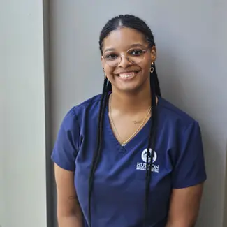 Brianna Waterton, wearing navy blue medical scrubs and glasses, poses for a professional portrait at Hudson Animal Hospital.