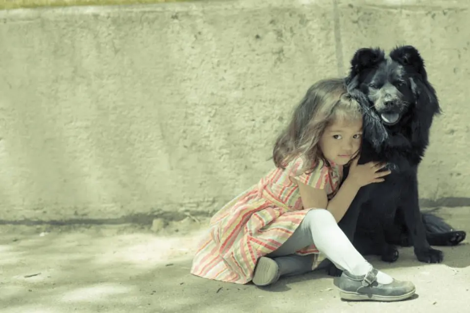 A young girl in a striped dress sitting on the ground and hugging a large black dog.