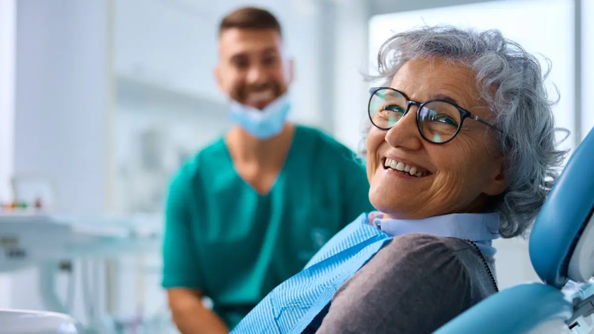 Senior female patient smiling in dental chair with dentist in background