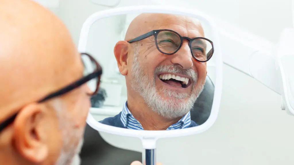 Senior male patient smiling while looking in mirror after getting dentures