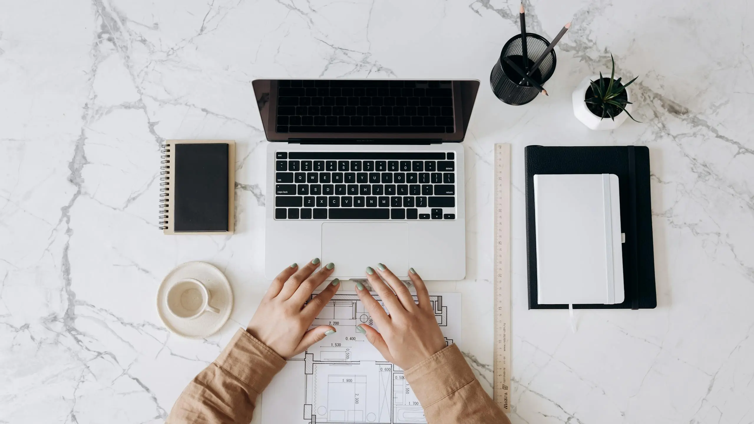 top view of a stylish home office desk with a laptop planner and coffee cup showing hands on a blueprint. 6894103 scaled