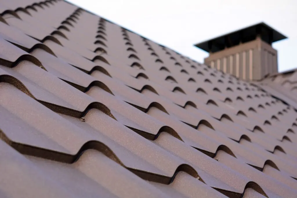 House with metal galvanized coating, chimney pipe, mansard windows and snow holding cornice on roof slope against sunset sky. Element of modern residential building with zinc rooftop sheets