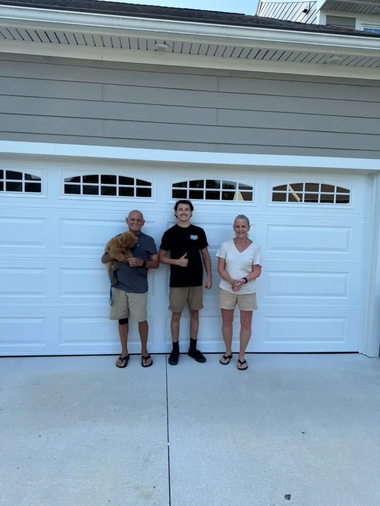 Three people and a brown dog standing in front of a white garage door on a driveway