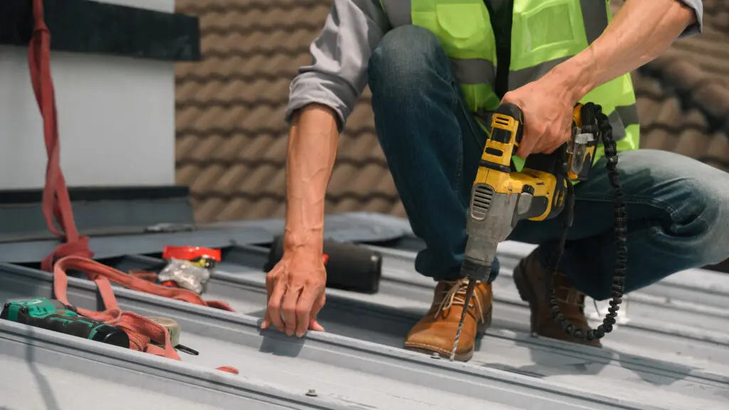 metal roof maintenance checklist Close up of a professional roofer in a safety vest drills into a metal roof panel during maintenance work, with tools and harness visible nearby.
