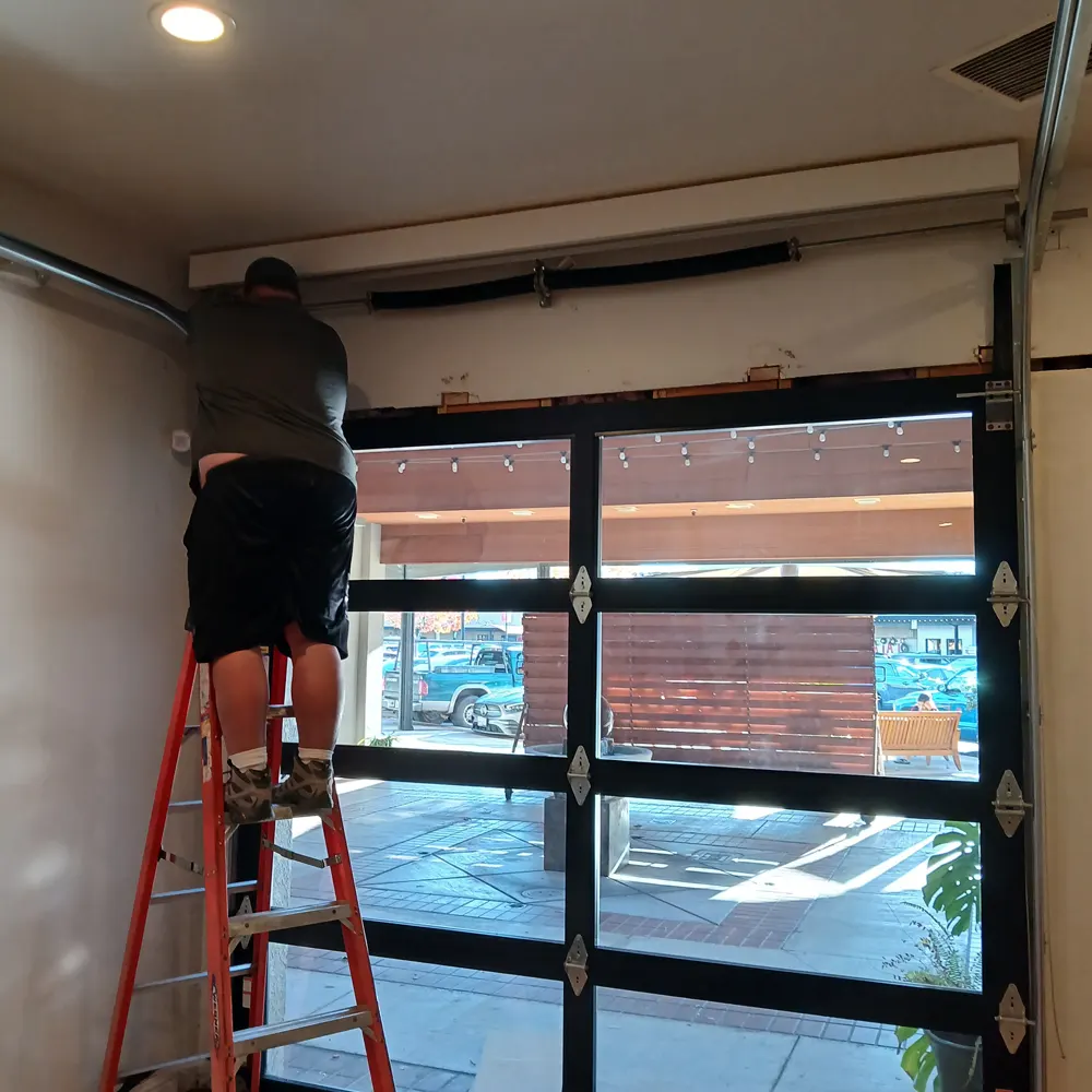 Technician on a ladder adjusting the hardware above a full-view glass garage door