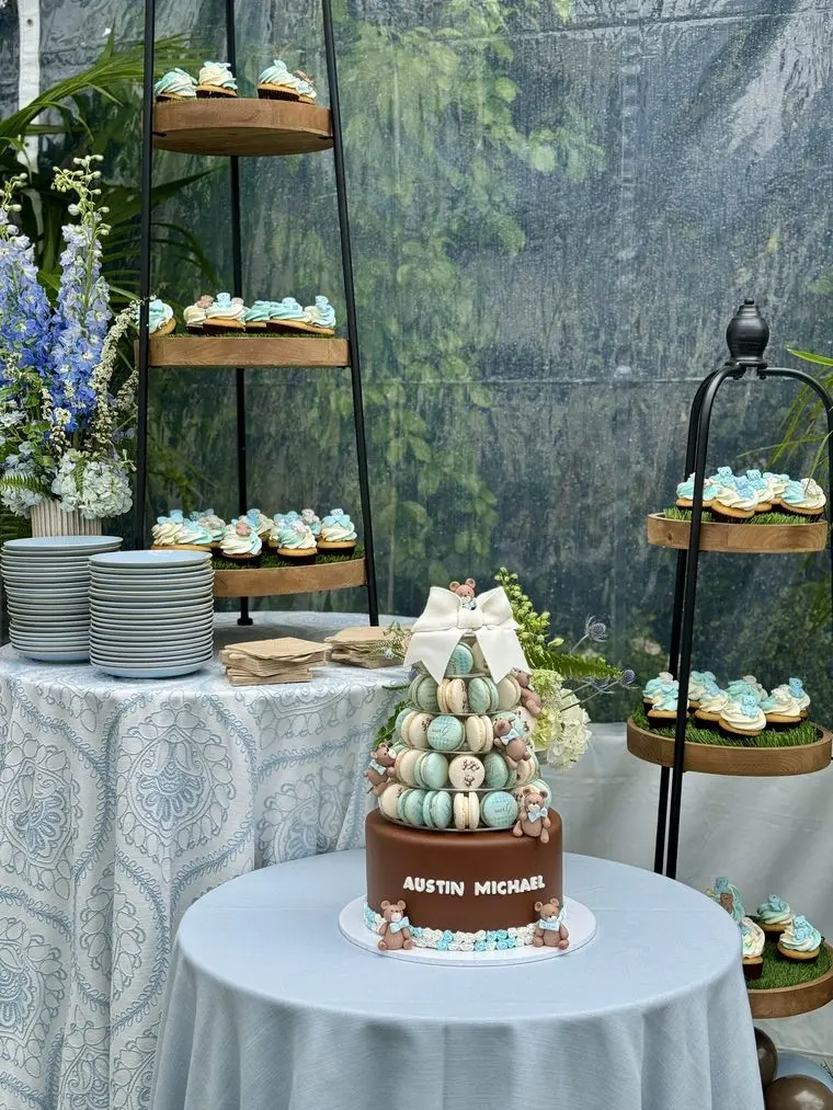Handheld Catering baby shower dessert table with tiered stands of decorated cupcakes and cookies beside a macaron tower on a linen-draped table