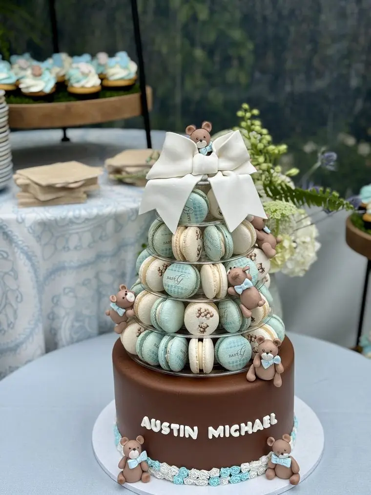 Handheld Catering baby shower macaron tower in soft blue and cream topped with a bow, set on a chocolate tier trimmed with sugar teddy bears