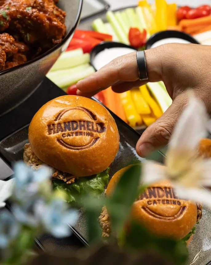 Hand holding a Handheld Catering branded slider bun with crudites and wings in background