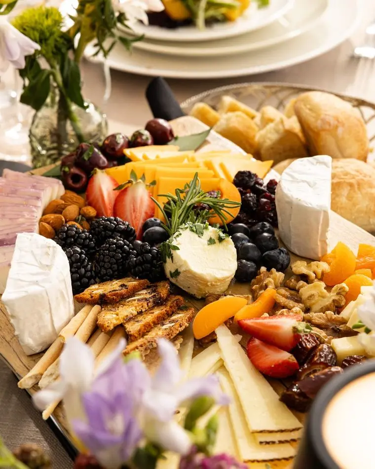Grazing board at a seated Handheld Catering event with brie, cheddar, manchego, herbed goat cheese, blackberries, strawberries, walnuts, dried apricots, dates and crostini, with place settings and flowers behind