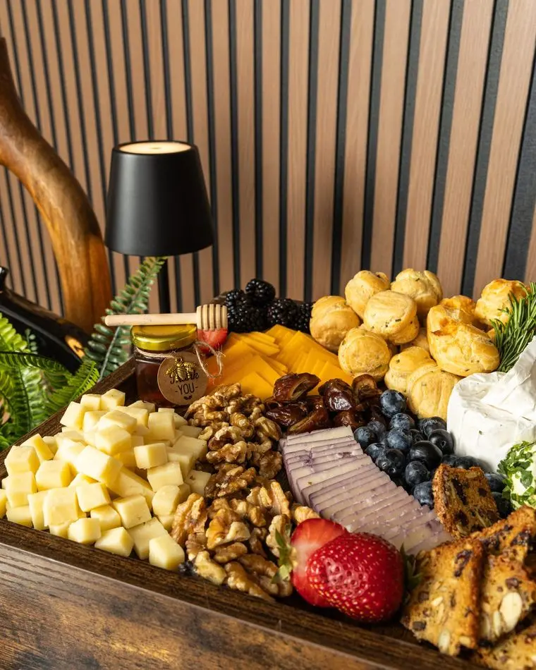 Handheld Catering cheese and charcuterie tray with gougeres, cubed cheddar, walnuts, dates, blueberries, blackberries, brie and a jar of local honey against a wood slat wall